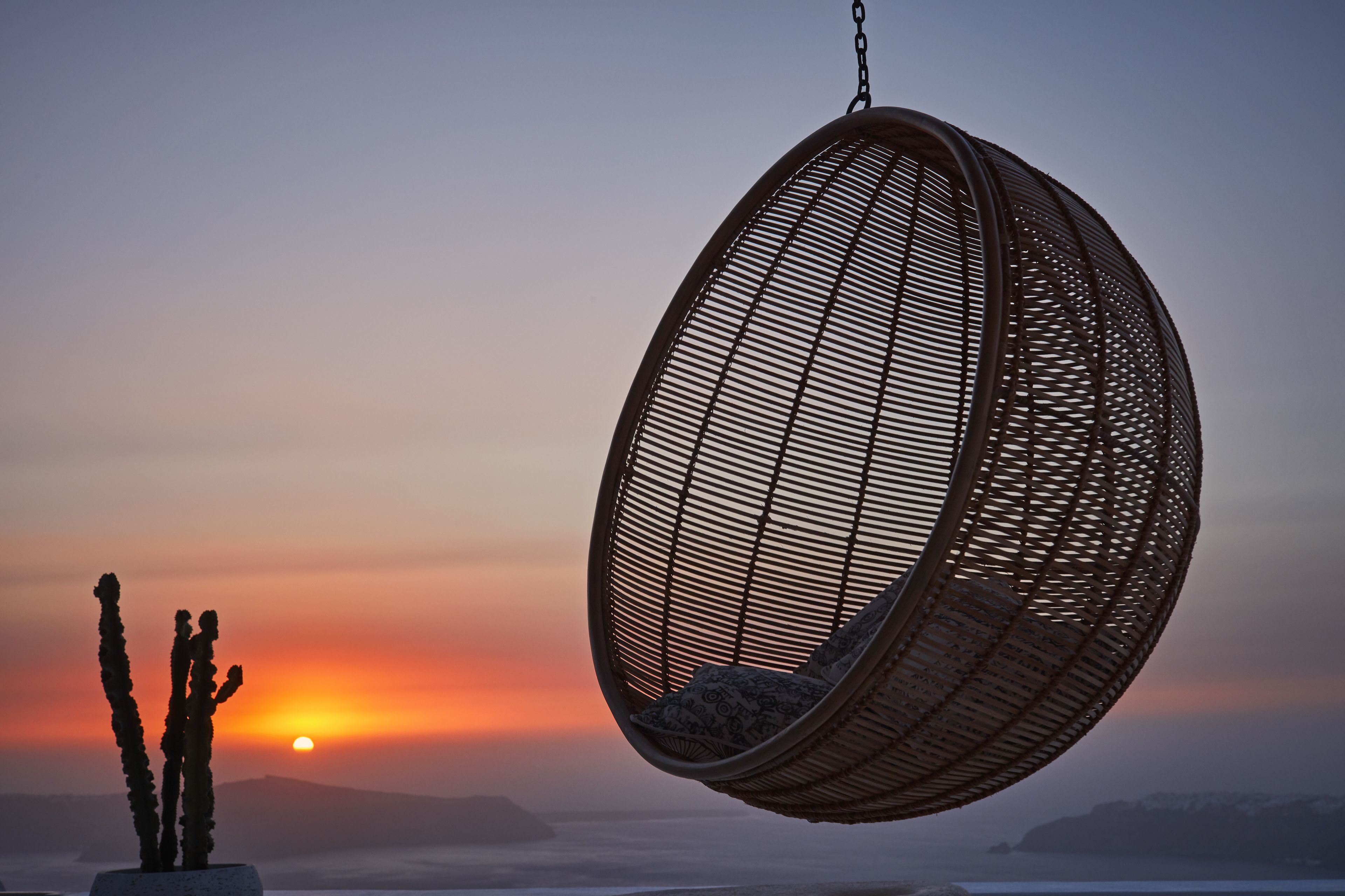 A hanging chair with a woven design, a cactus, and a sunset in the background.