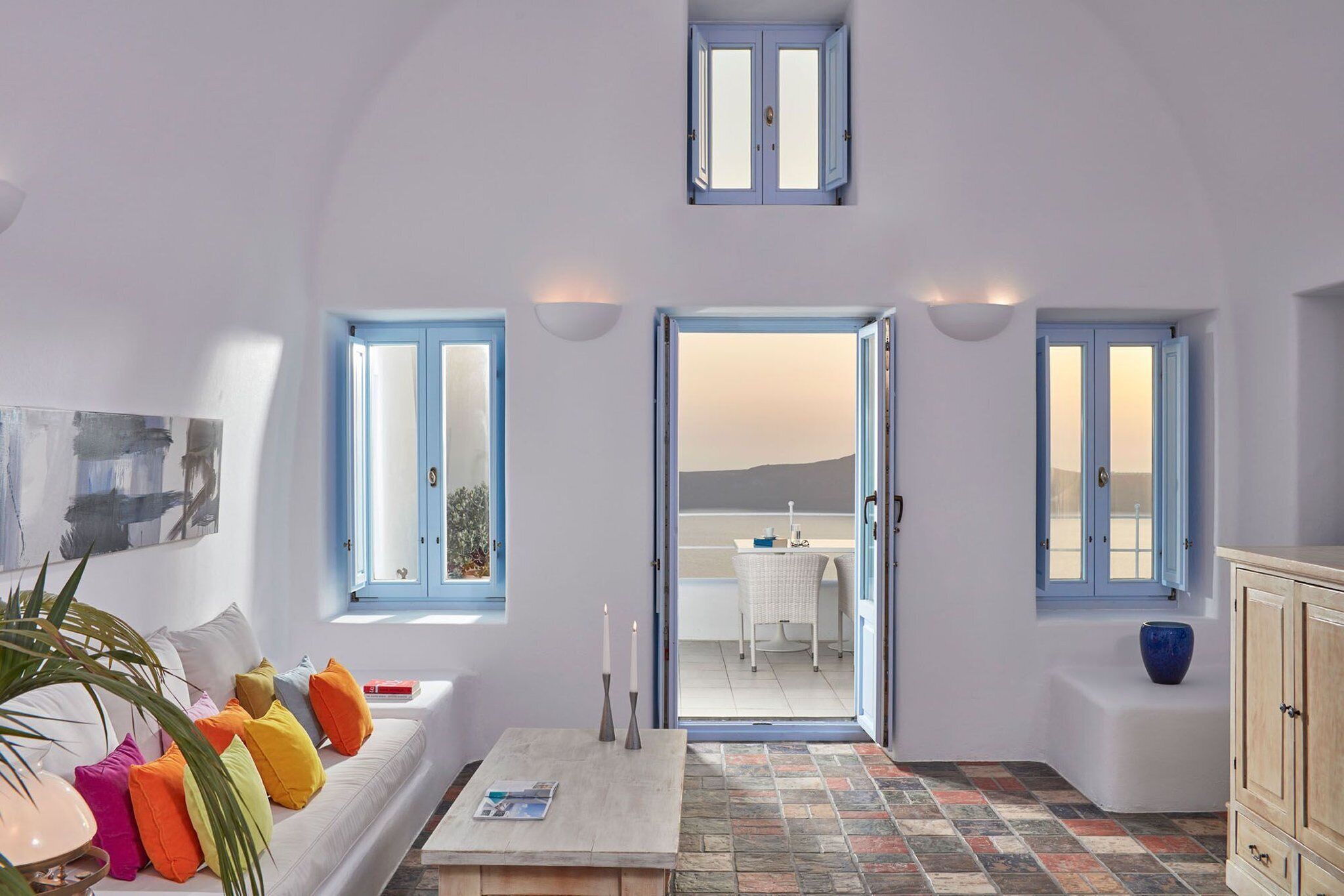 Una habitación con un sofá blanco, almohadones coloridos, una mesa con una revista y vistas a un balcón con mesa y sillas.
