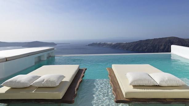 A infinity pool with two lounge chairs, each with a pillow, overlooking a coastal landscape with cliffs and the sea. - Astra Suites (Santorini)