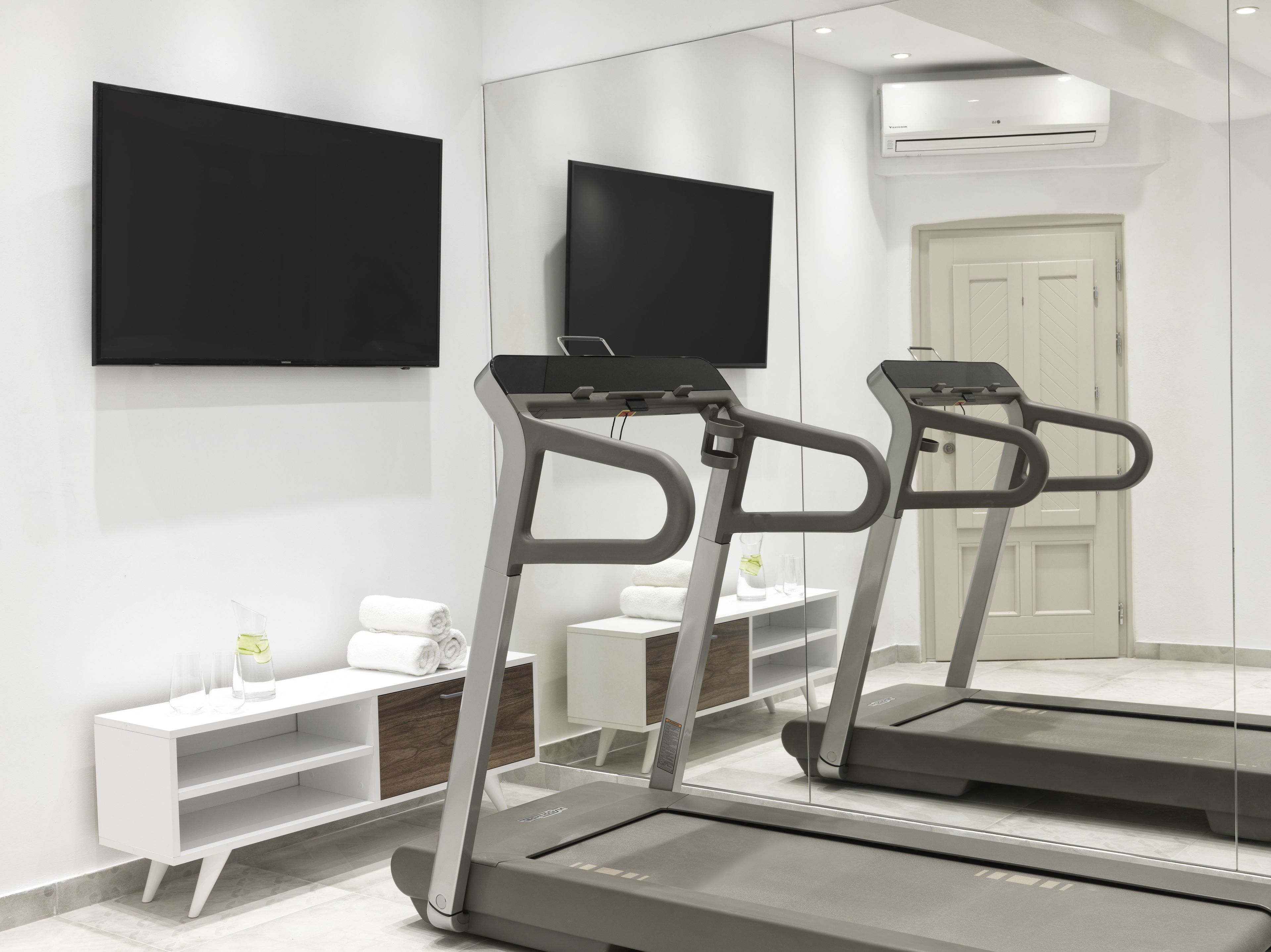 A modern hotel room with a treadmill, a flat-screen TV, a white vanity, and a large mirror.