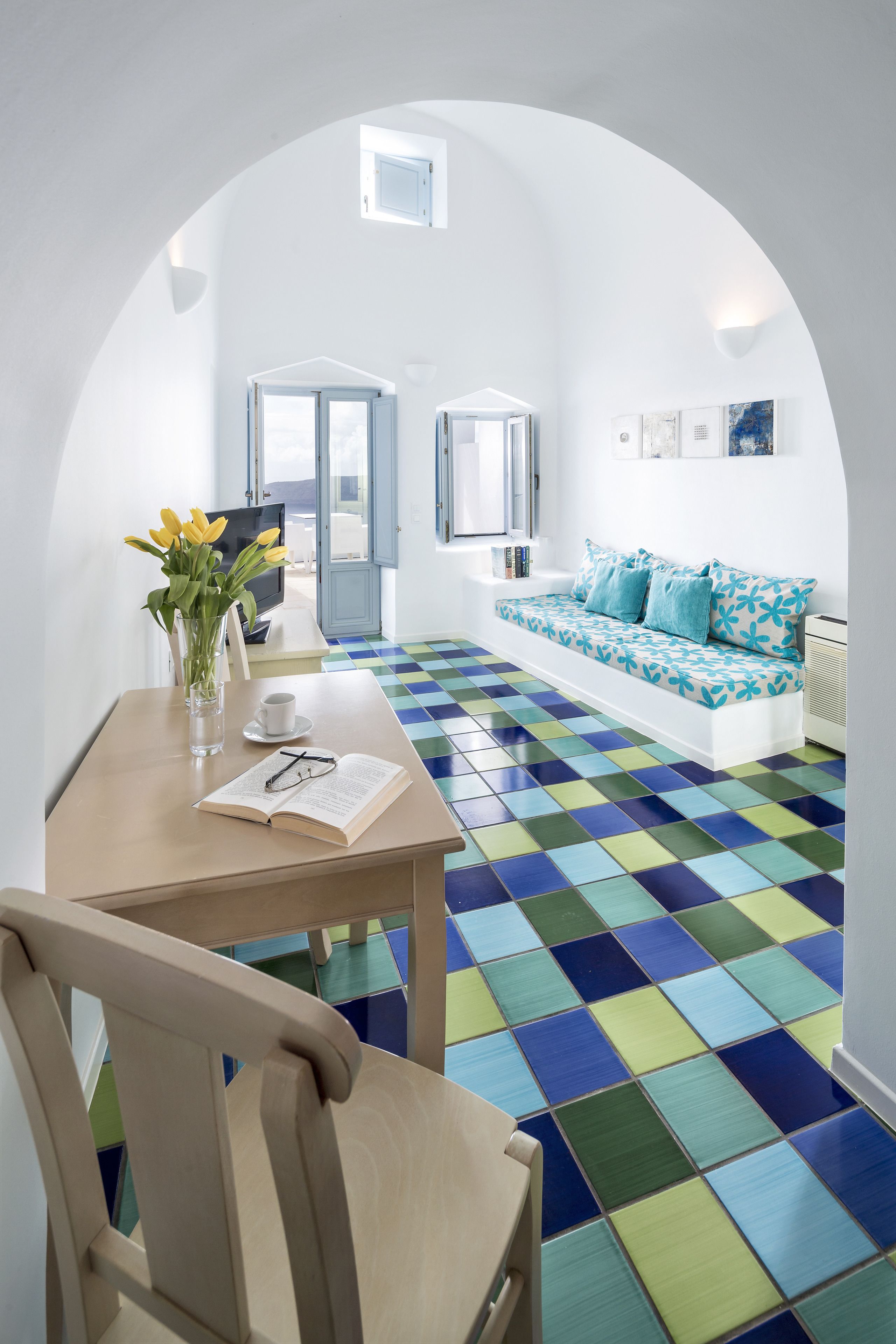 A modern room with a checkered floor, a wooden table with a book and glasses, a sofa with patterned cushions, and a TV.