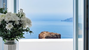 A table with a vase of white flowers, a book, and an apple, positioned by a window with a view of the sea and a rock. - Astra Suites (Santorini)
