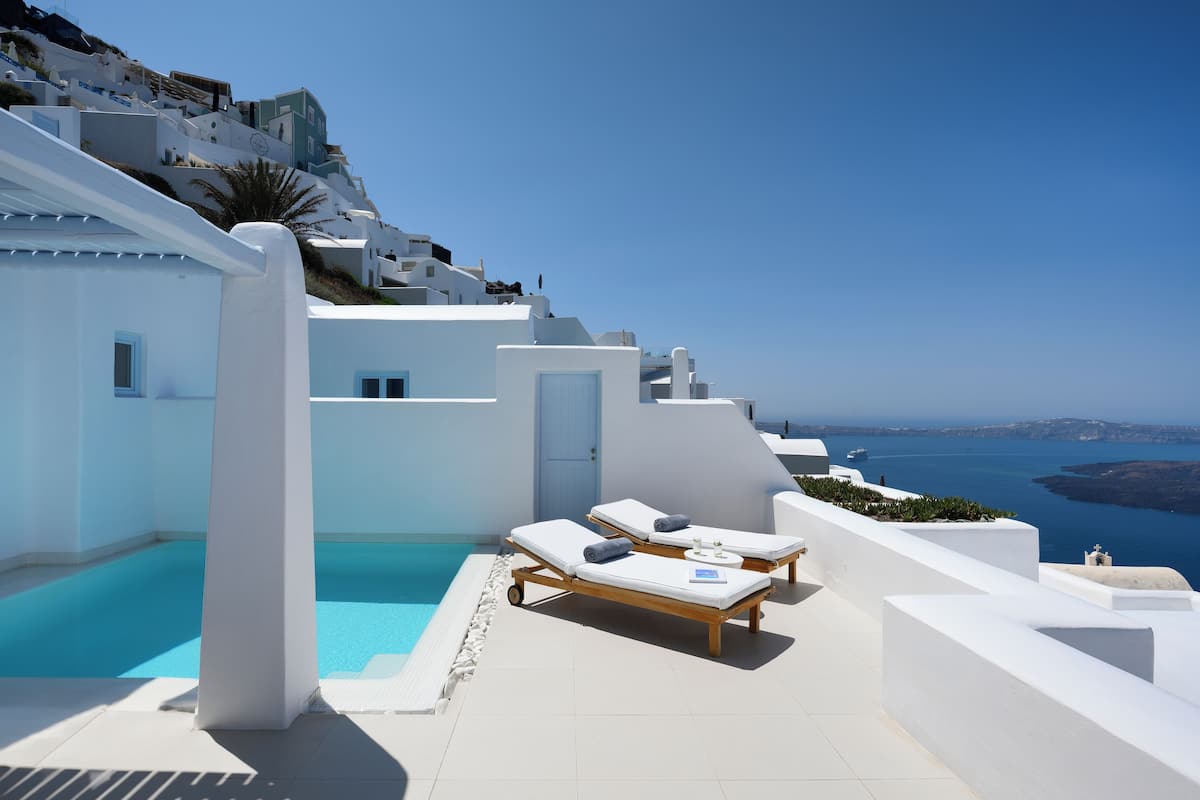 A white-walled building with a pool and lounge chairs overlooking a clear blue sea.