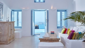 A spacious room with a white sofa, a wooden table, and a view of the sea through open doors. - Astra Suites (Santorini)