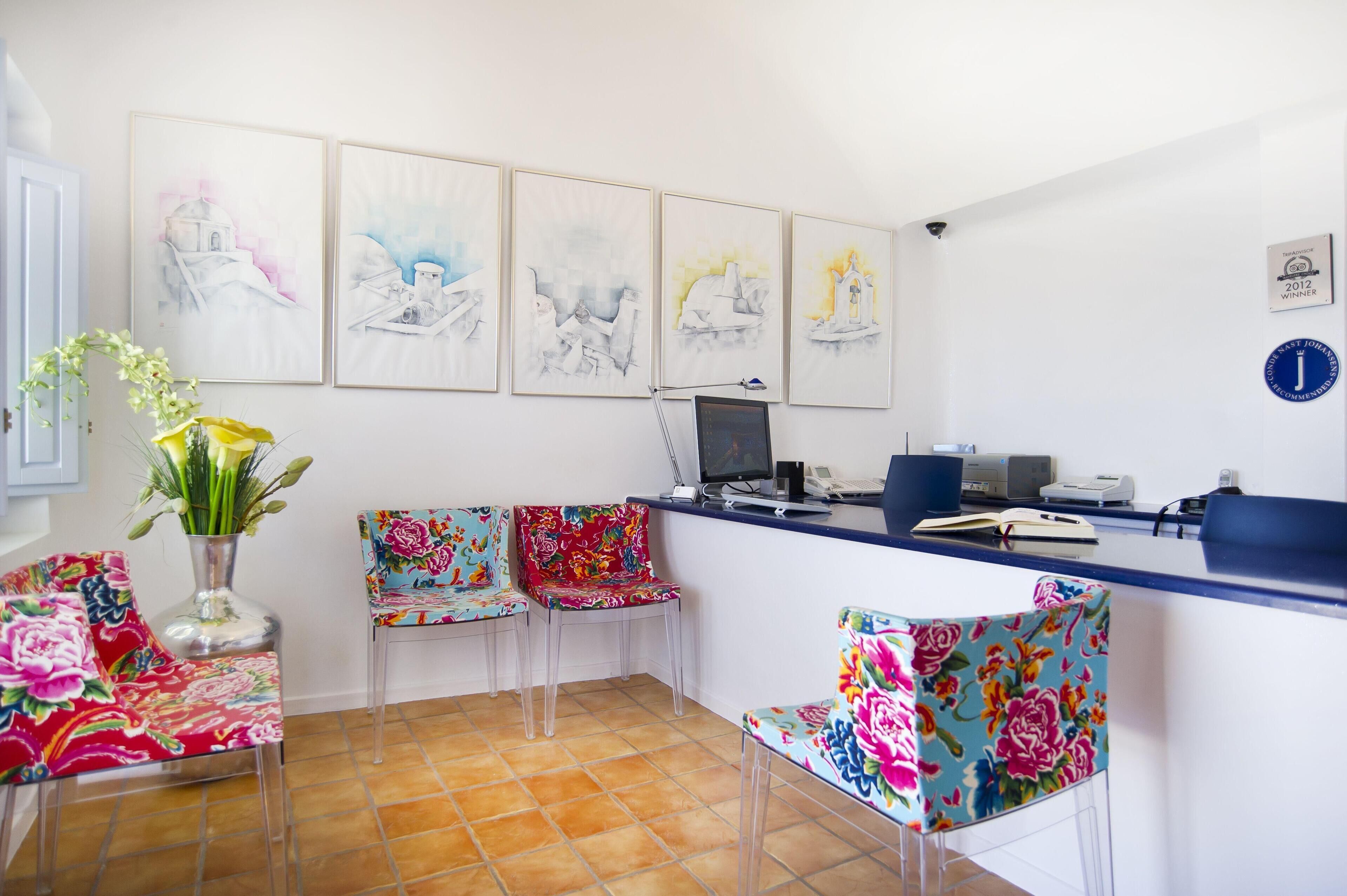 Zona de recepción con un escritorio, computadora, impresora y dos sillas. Cuatro obras de arte enmarcadas adornan la pared, y un jarrón con flores se encuentra a la izquierda.