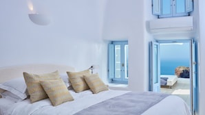 A bedroom with a bed, pillows, a nightstand, and a view of the sea through an open door. - Astra Suites (Santorini)