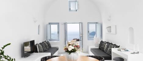 A modern, minimalist room with a round wooden table, black chairs, and a white sofa. There are three windows providing views of the sea.