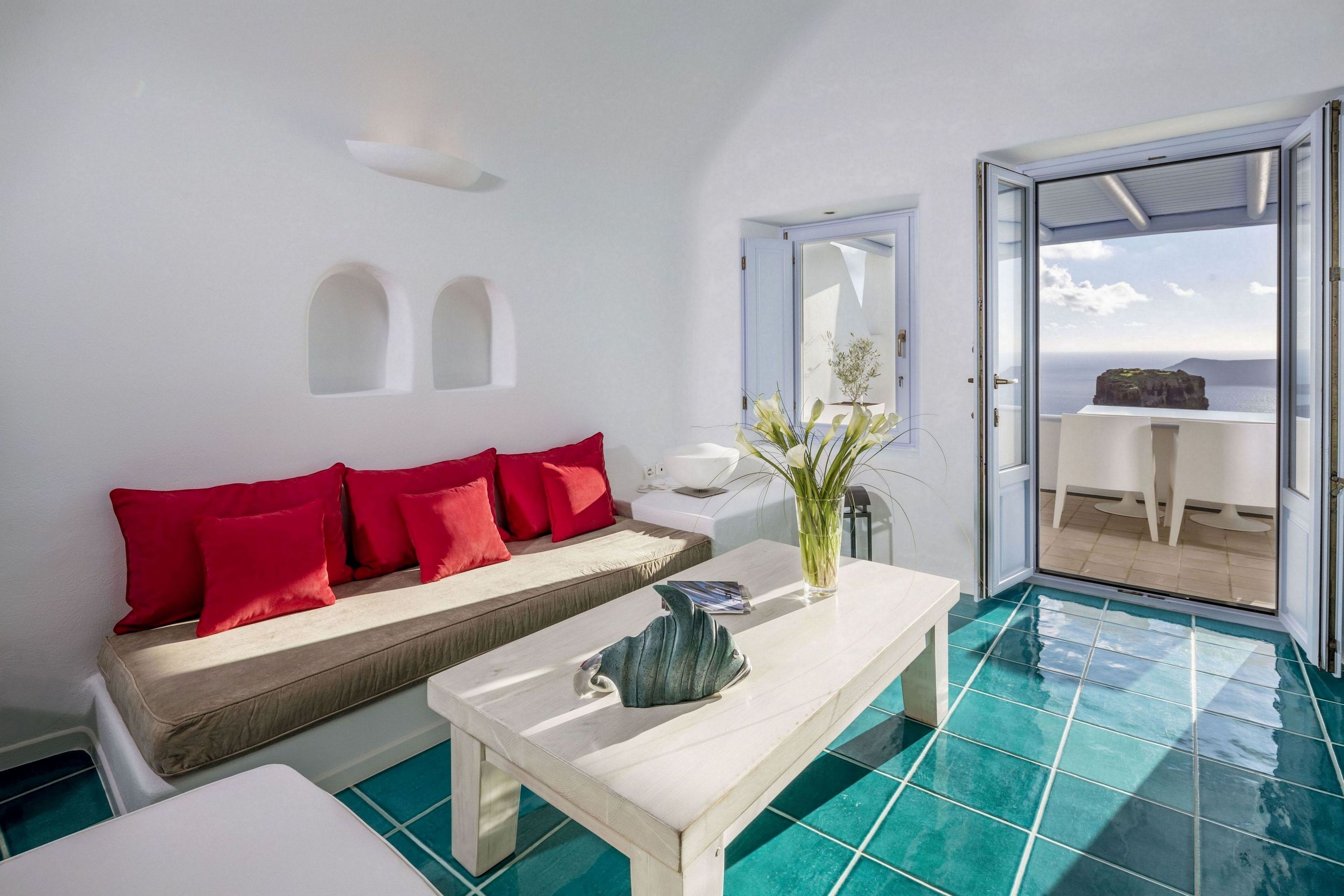 Una habitación con un sofá blanco, cojines rojos, una mesa blanca y vistas a un balcón con mesa y sillas desde una puerta abierta.