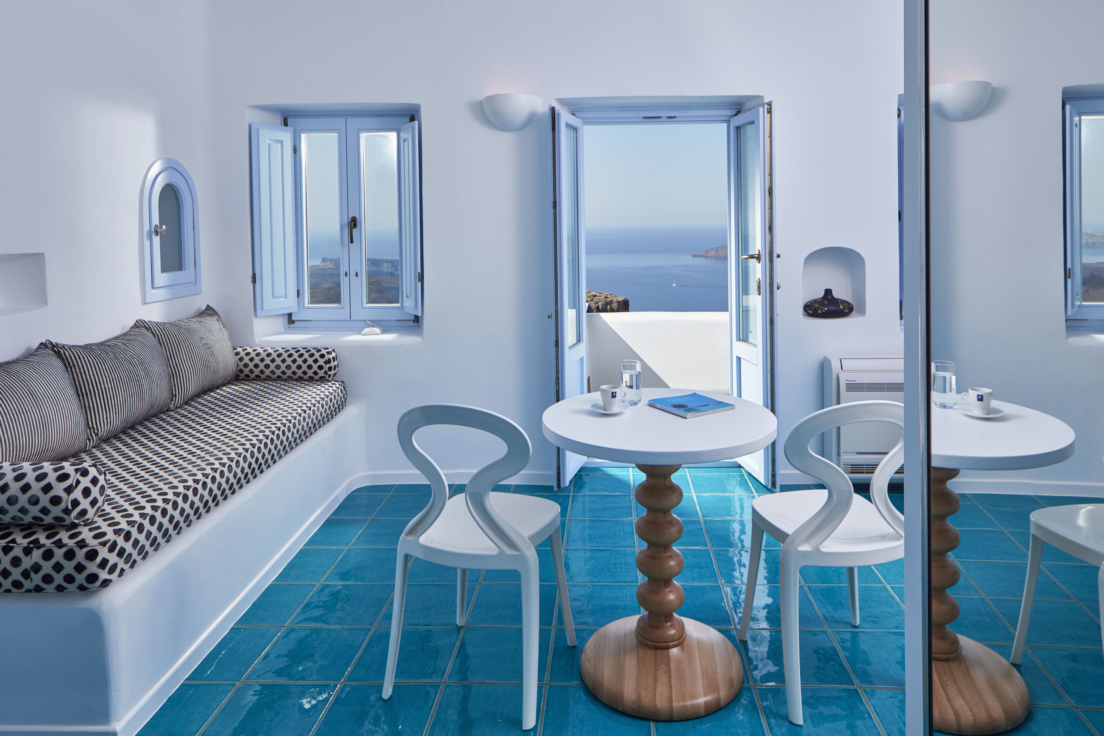 A modern room with a white sofa, two white chairs, a round wooden table, and a view of the sea through open doors.