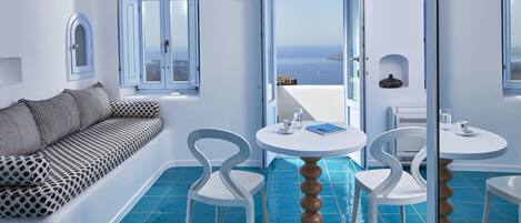 A modern room with a white sofa, two white chairs, a round wooden table, and a view of the sea through open doors.