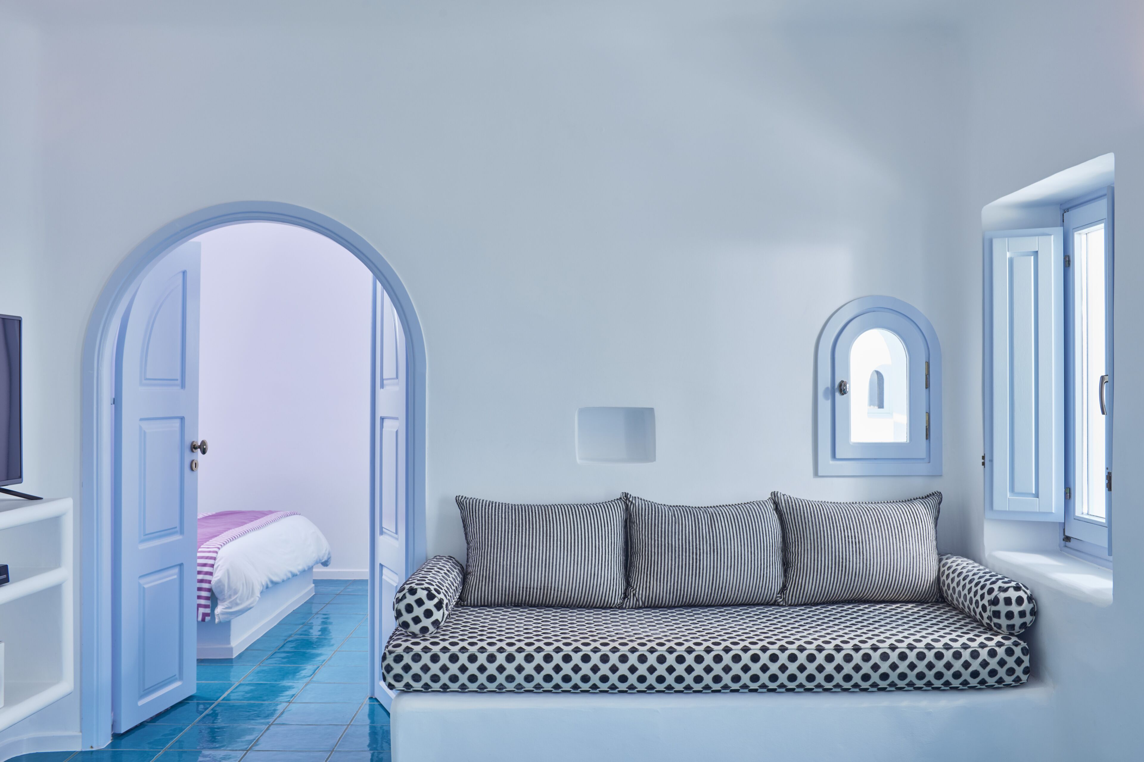 A room with a patterned sofa, a bed with a striped blanket, and a TV on a white shelf.