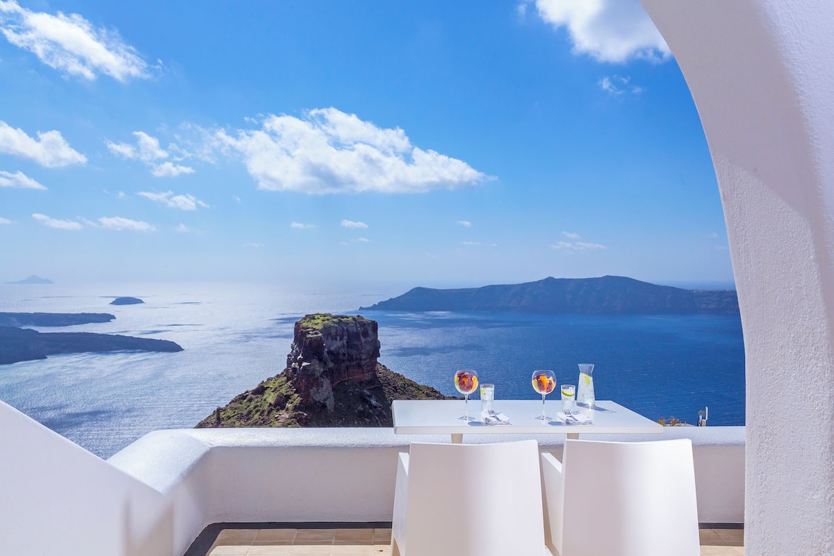 A table set with two glasses of water and a bottle, overlooking a scenic view of the sea and islands.