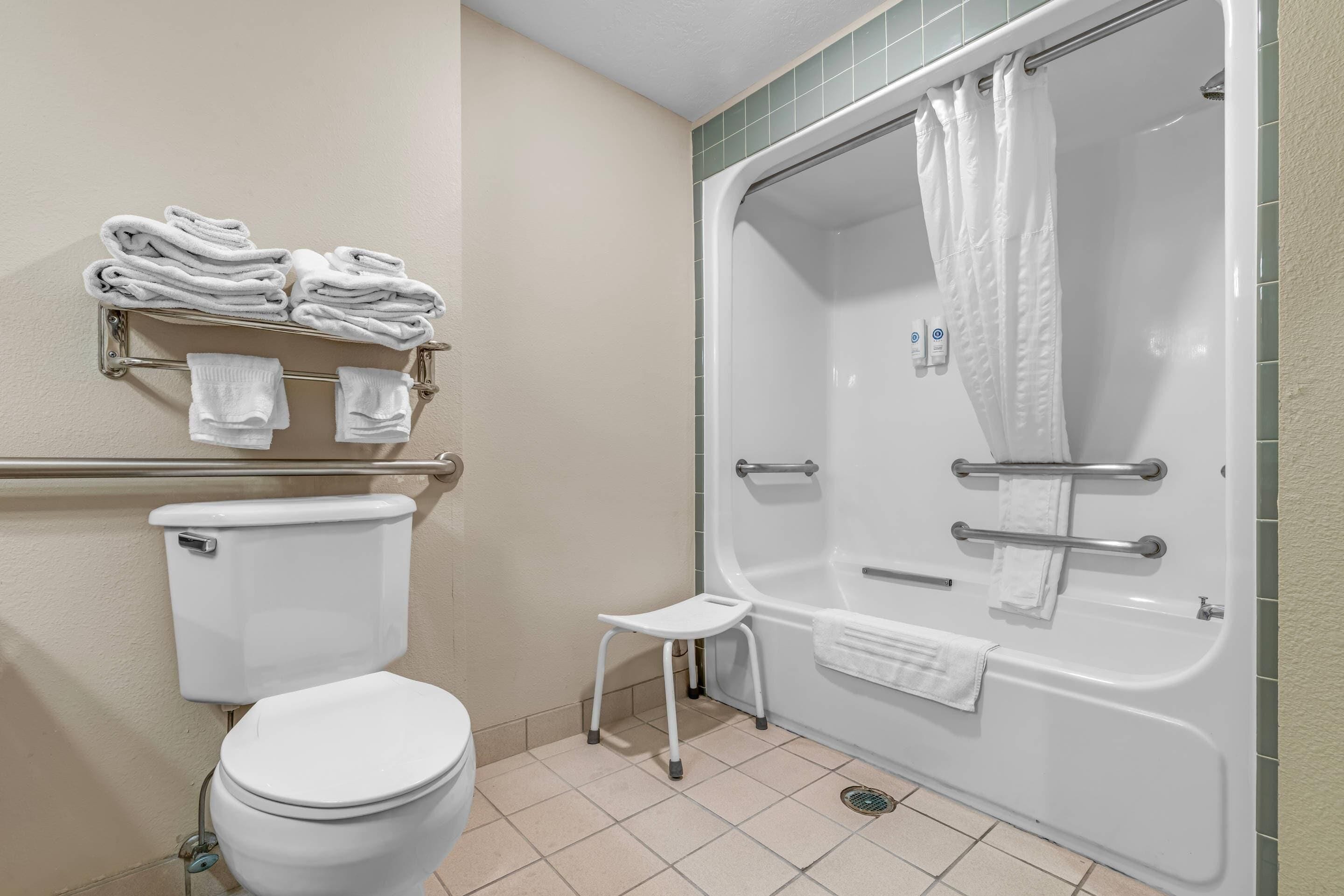 Un baño con inodoro, ducha con puerta de vidrio, un taburete pequeño y un perchero con toallas dobladas.