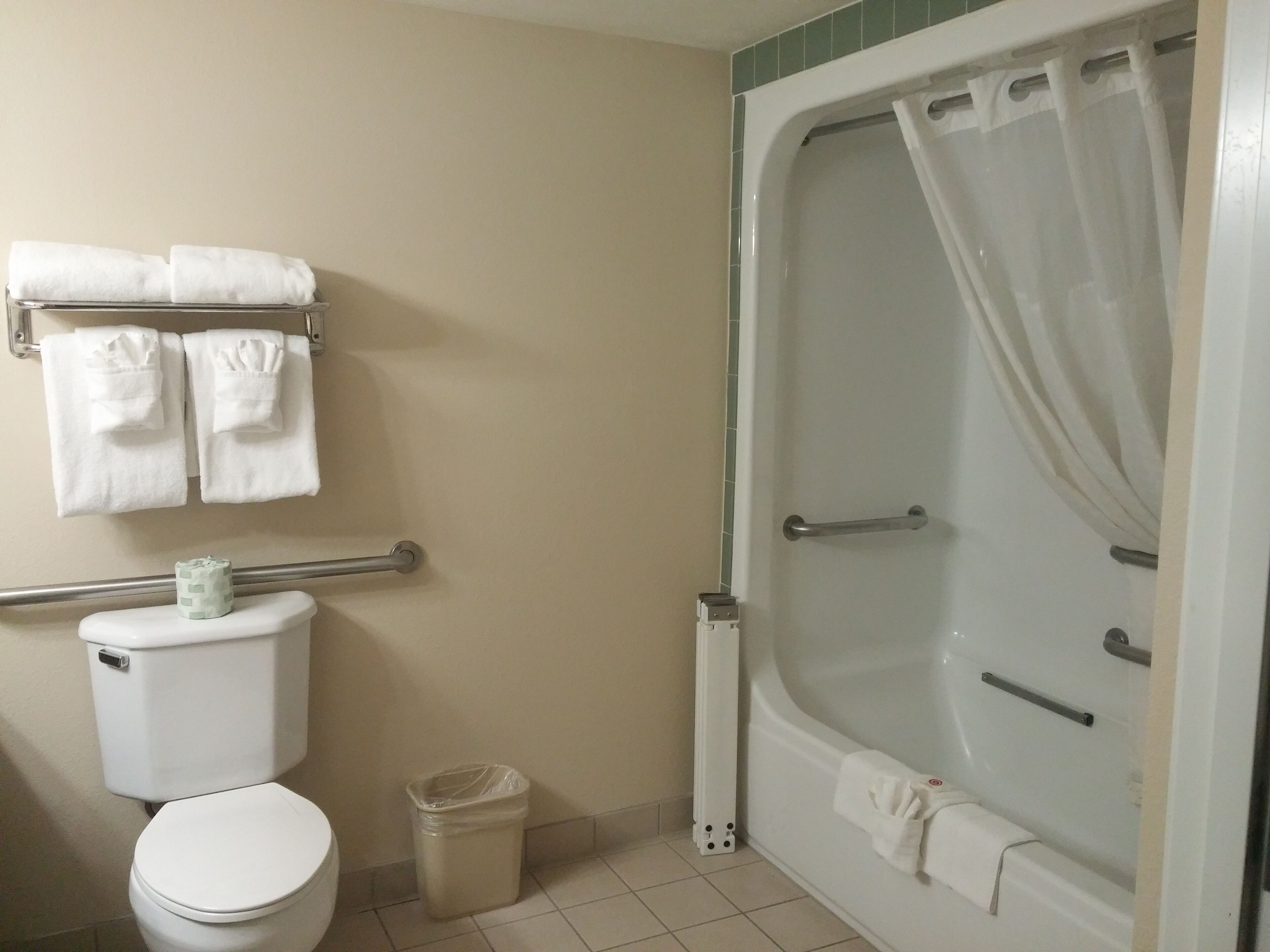 A bathroom with a toilet, a trash can, a towel rack with towels, and a bathtub with a shower curtain.