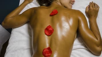 A person lying on a bed with a white sheet and pillow, receiving a back massage with red rose petals.