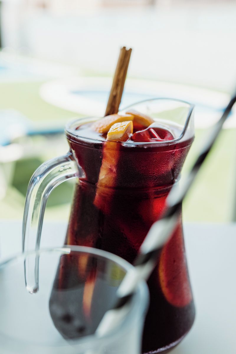 A glass pitcher with a straw and a cinnamon stick, containing a dark beverage with ice cubes.