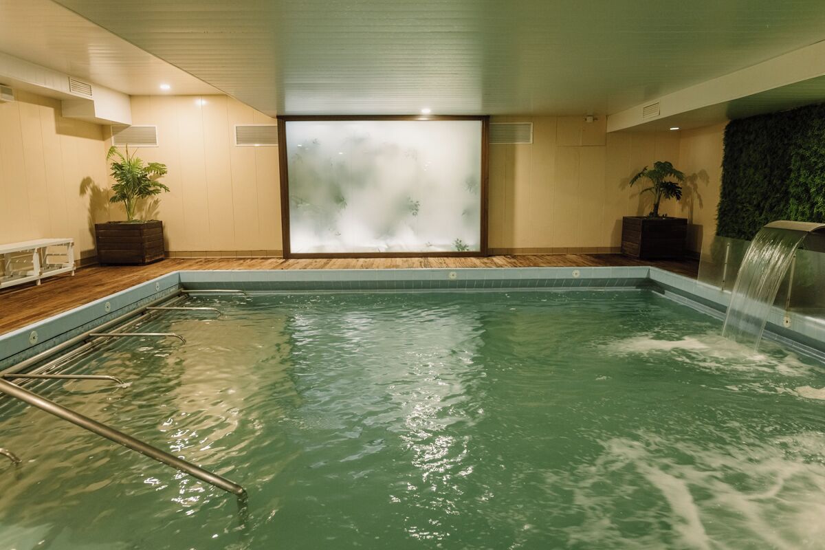A large indoor swimming pool with a wooden floor, a glass wall, and a waterfall feature.
