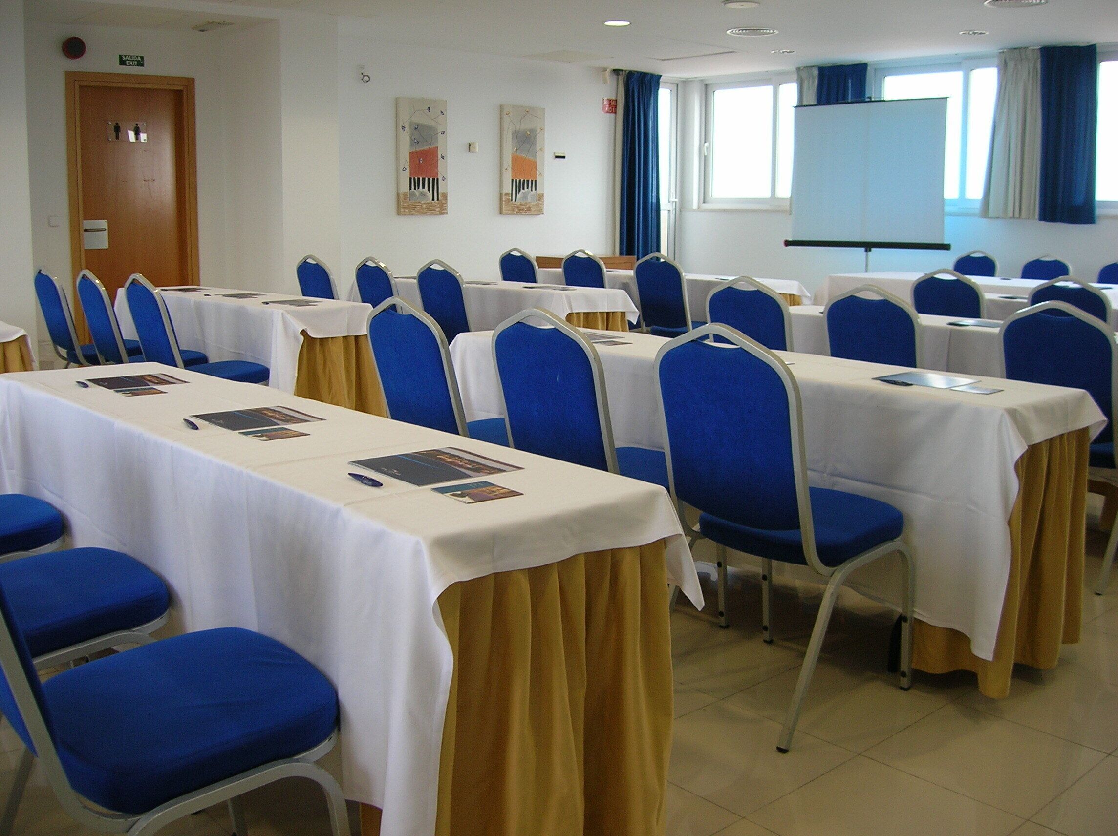 Uma sala de conferência com mesas e cadeiras dispostas em fileiras, uma lousa branca e uma tela de projetor.
