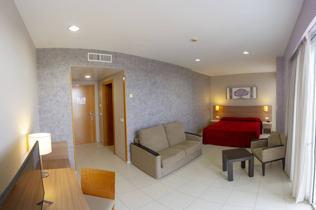A hotel room with a sofa, armchair, small table, and a red bed. There is a door leading to another room and a window with curtains.
