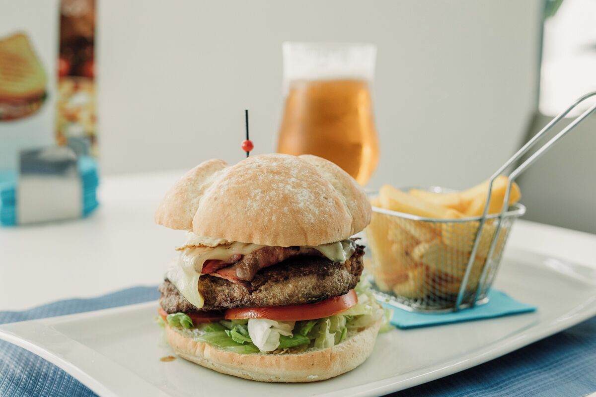 Una hamburguesa con un palillo encima, un vaso de cerveza y una cesta de papas fritas sobre un plato blanco.