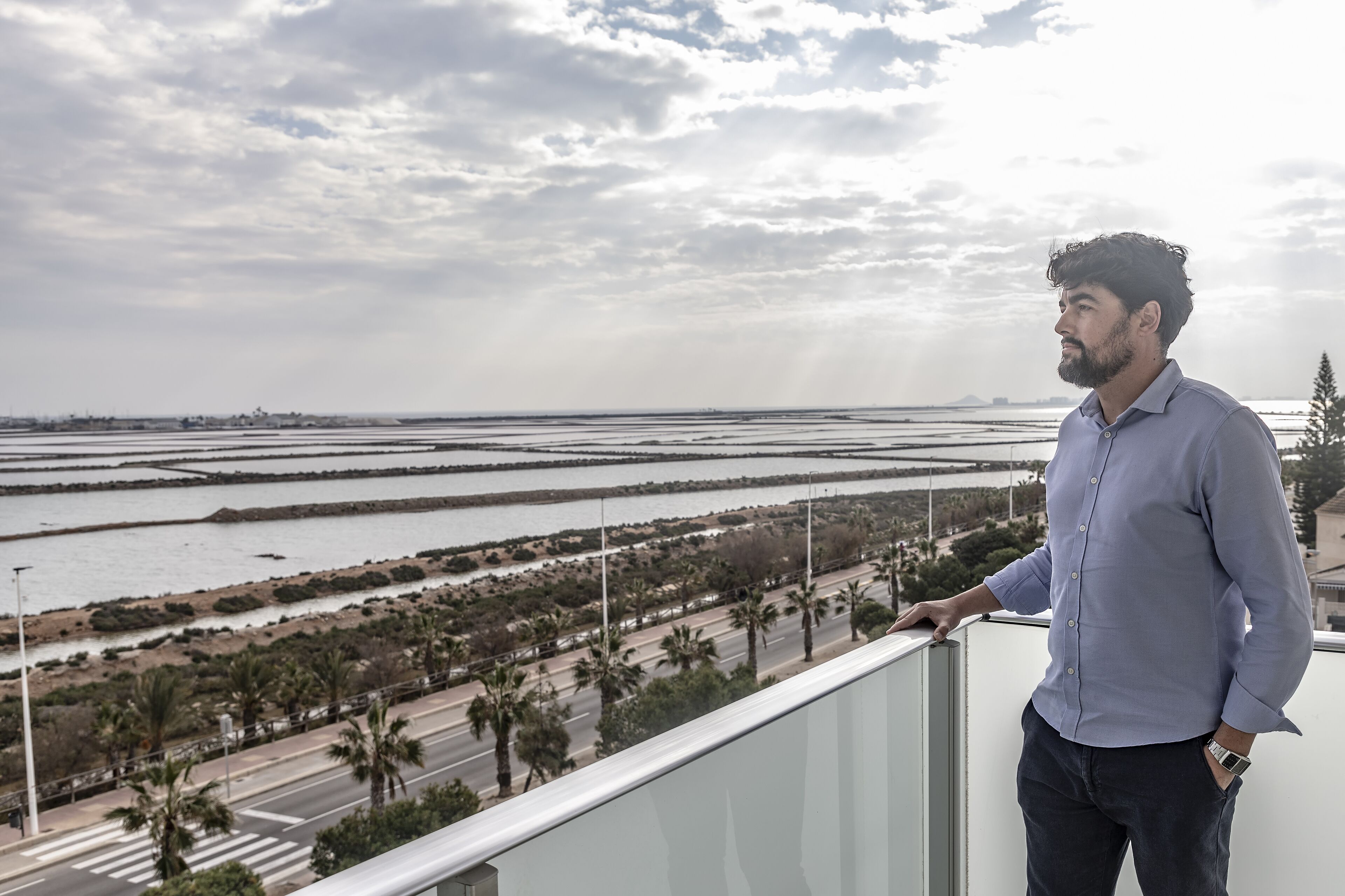 Un uomo in piedi su un balcone con vista su un paesaggio costiero, una strada, palme e una lontana vista del mare.