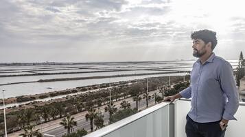 Un uomo in piedi su un balcone con vista su un paesaggio costiero, una strada, palme e una lontana vista del mare.
