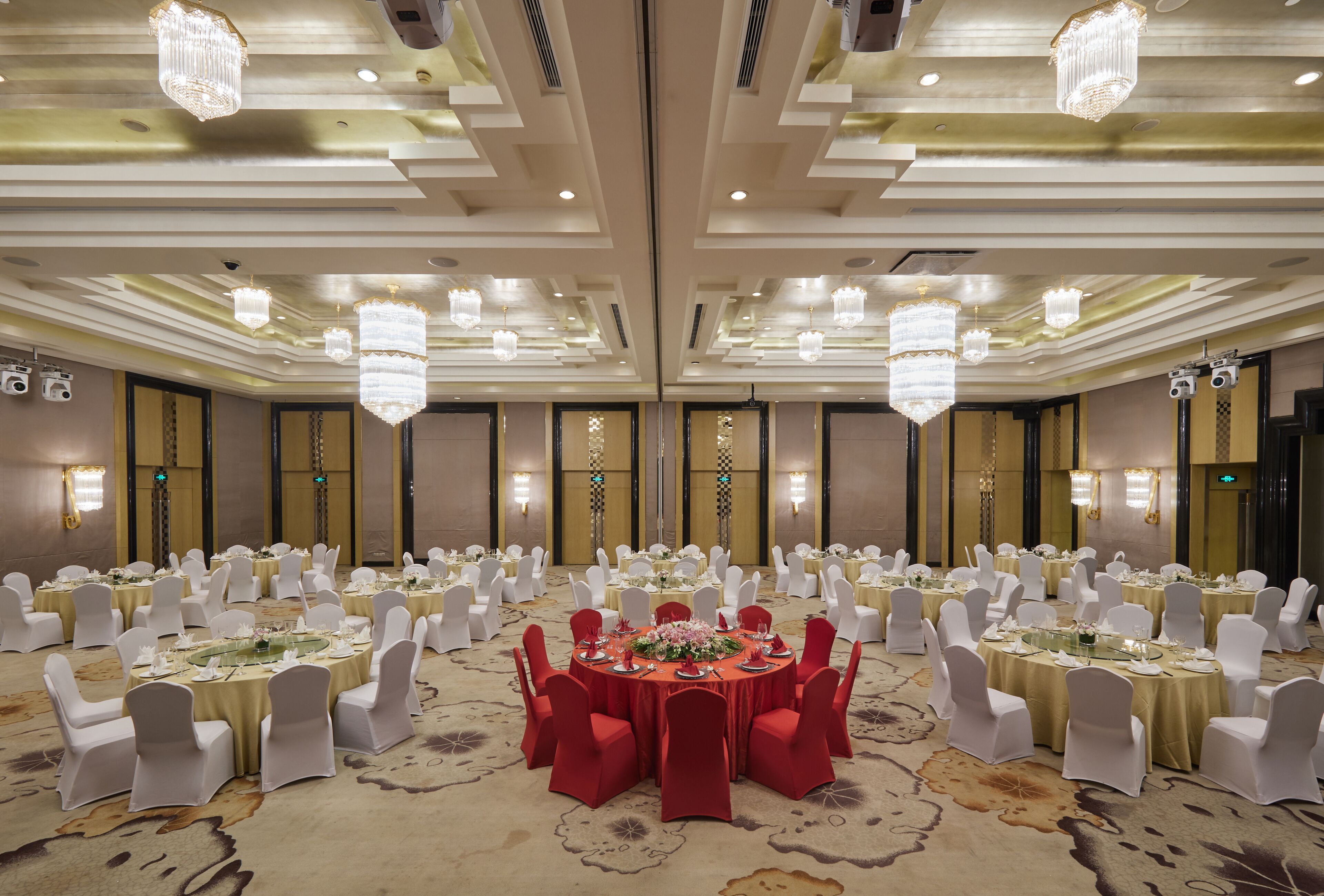 Un salón de banquetes con mesas redondas, sillas blancas y rojas, candelabros y suelo alfombrado.