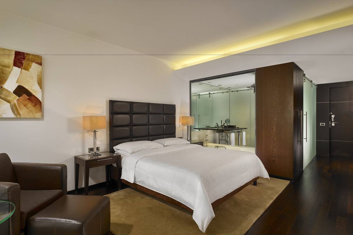 Ein modernes Hotelzimmer mit einem großen Bett, einer Sitzecke mit Glastisch, einem Gemälde an der Wand und einem durch eine Glastür einsehbaren Badezimmer.