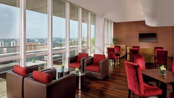 Un moderno vestíbulo de hotel con ventanas amplias, sofás de cuero, sillas rojas y suelo de madera.