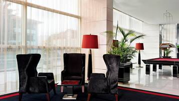 Um lobby moderno com duas poltronas pretas, um abajur vermelho, mesa de bilhar e uma janela grande com cortinas leves.