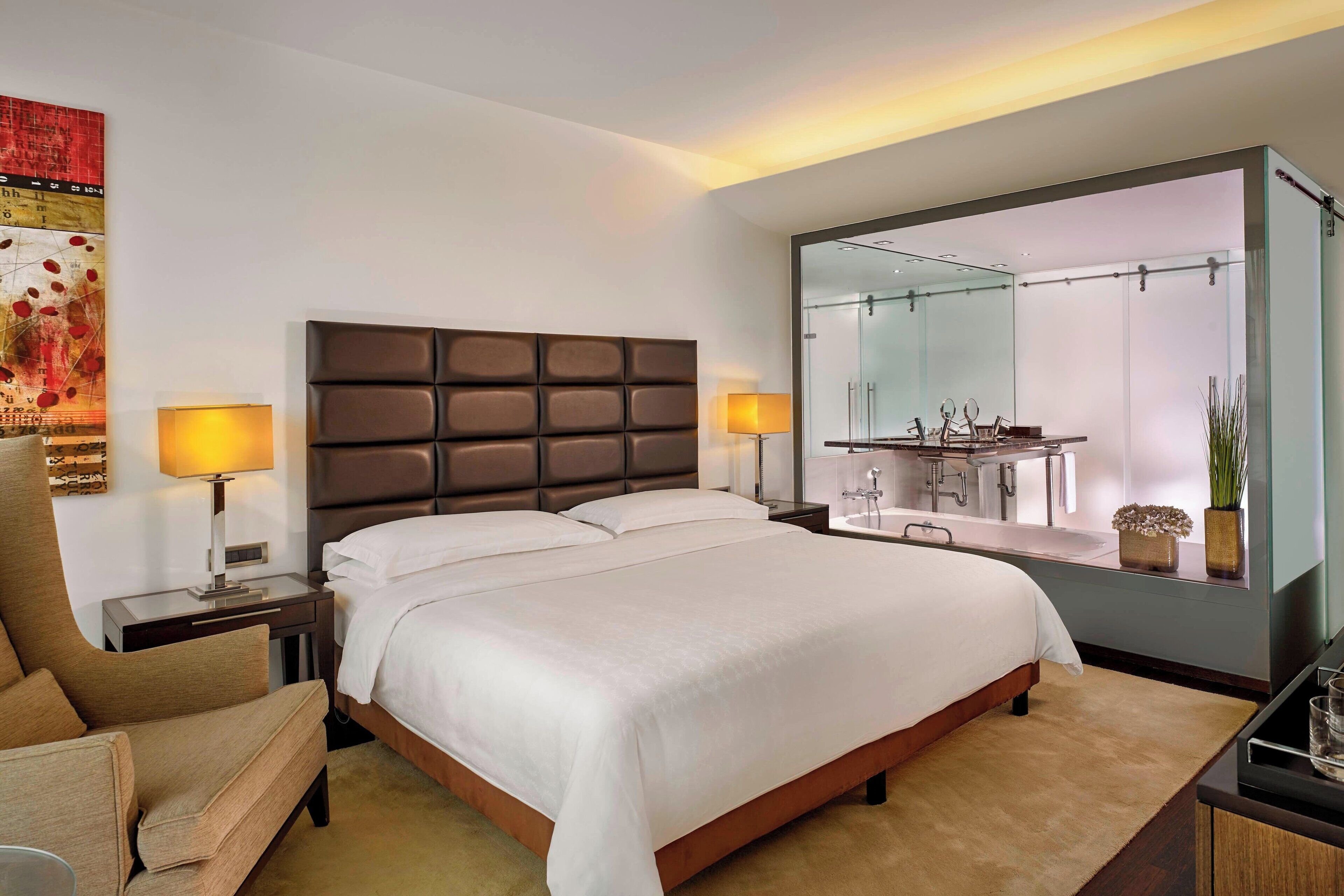 A modern hotel room with a large bed, a headboard with a padded design, two bedside tables with lamps, a large mirror, and a view of the bathroom.