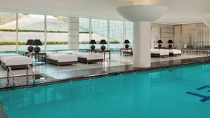 Un área de piscina interior moderna con sillones de descanso, una cama y ventanas amplias que ofrecen vistas a un jardín ajardinado.