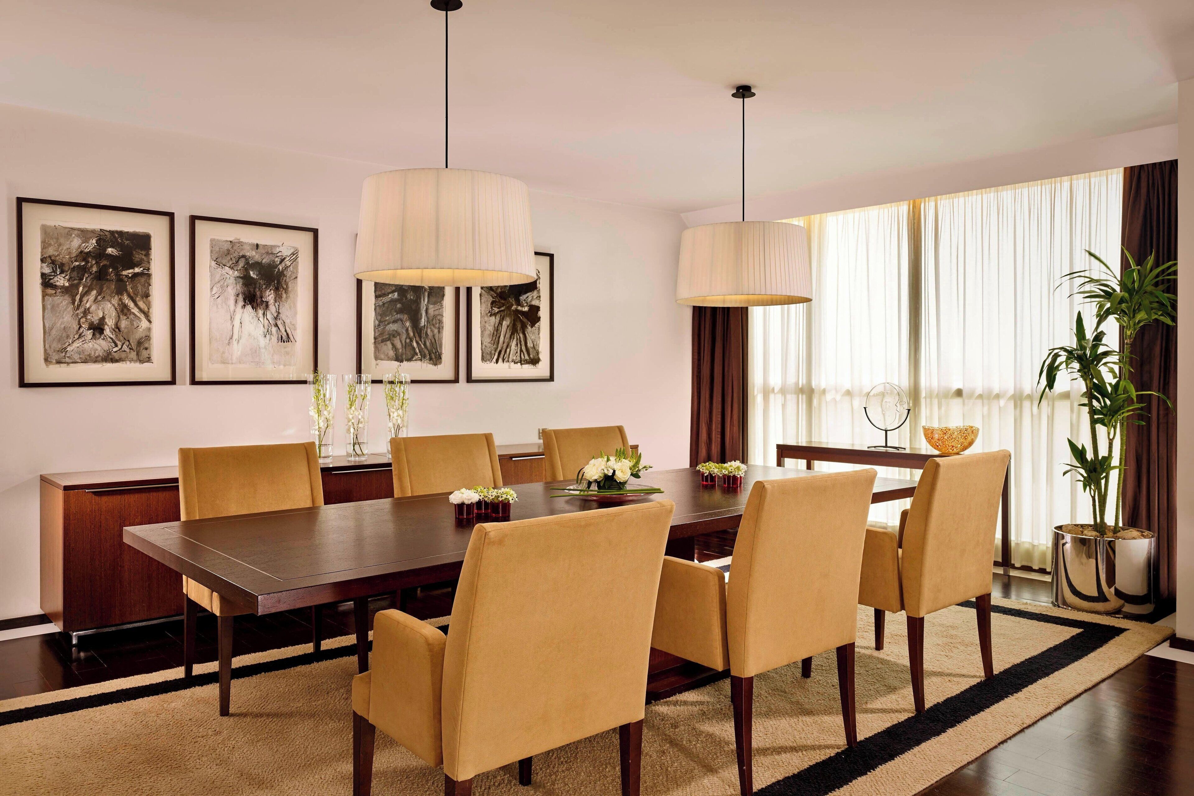 A dining room with a long table, six chairs, two hanging lamps, and framed artwork on the wall.