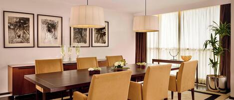 A dining room with a long table, six chairs, two hanging lamps, and framed artwork on the wall.
