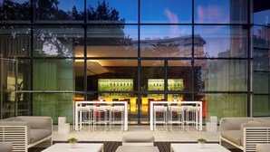 Un área de estar exterior moderna con mobiliario blanco y una terraza de madera, con vistas a un bar con iluminación verde en su interior.