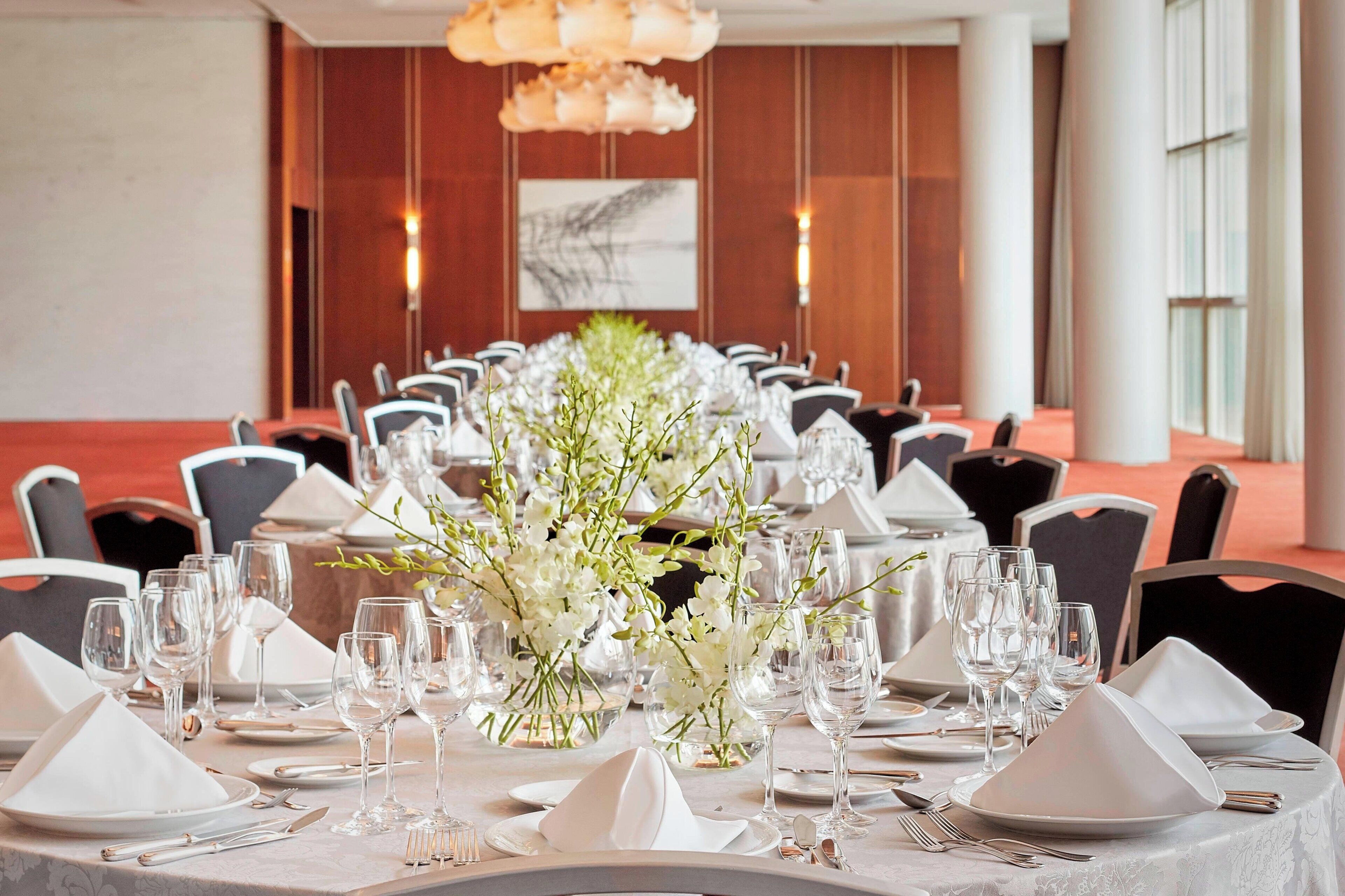 A long banquet table set with white tablecloths, silverware, and clear glasses, flanked by chairs with black cushions.
