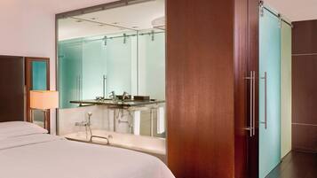 A modern hotel room with a large bed, a bathroom with a glass shower enclosure, and a wooden wardrobe.