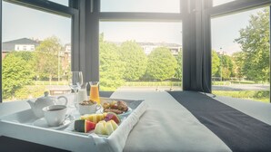 A breakfast tray with coffee, fruit, and pastries on a bed with a view of a park and buildings.