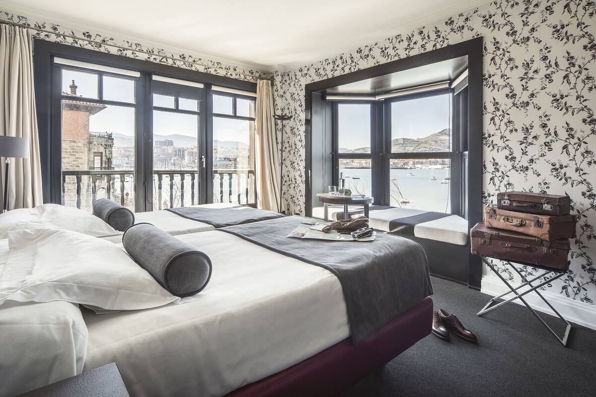 A hotel room with a large bed, a small table, a suitcase, and a view of the city and water.