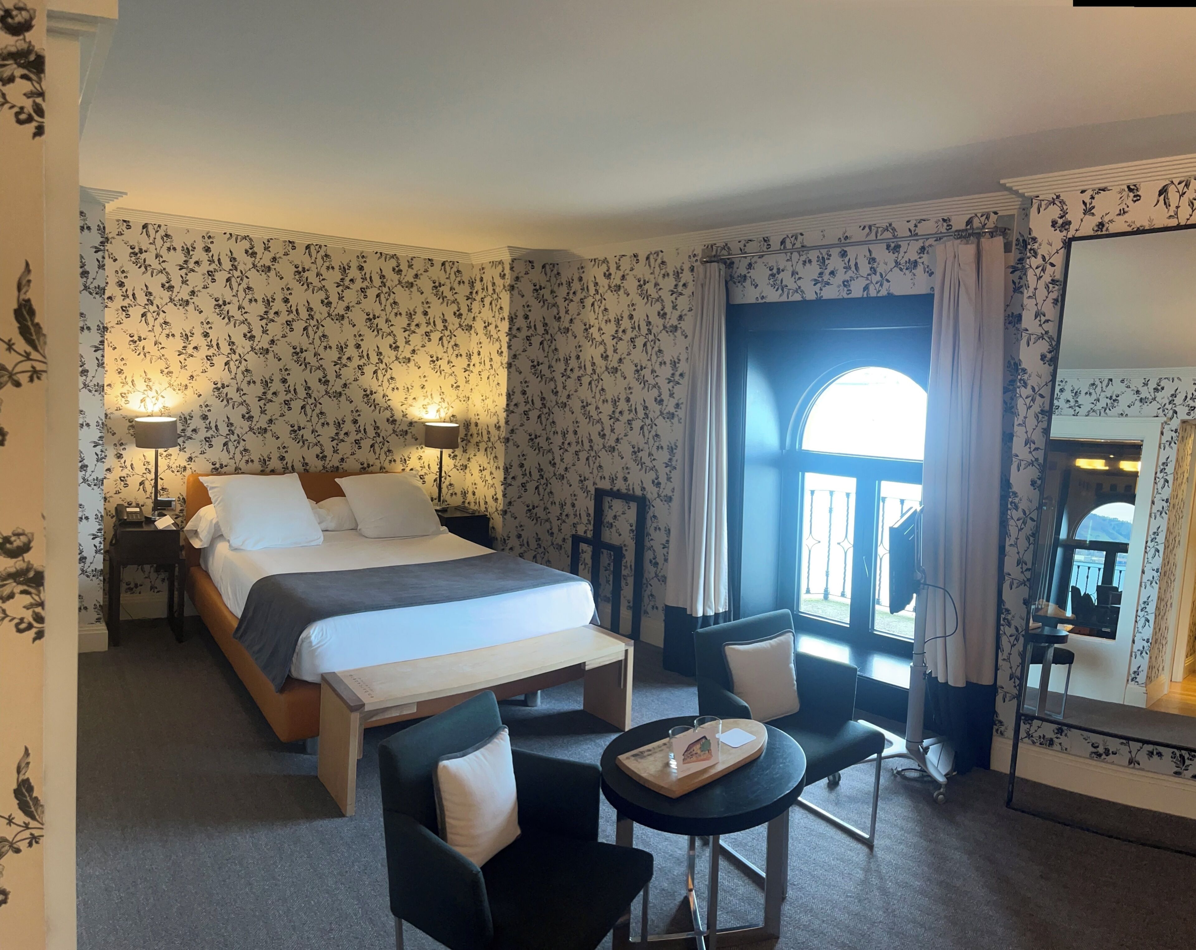 A hotel room with a bed, bedside lamps, a desk with a chair, a small table with a tray, and a large mirror.