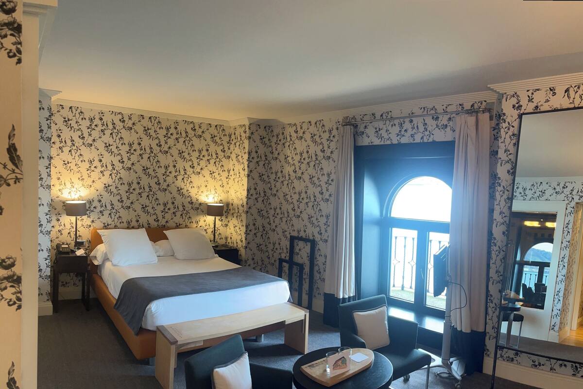 A hotel room with a bed, bedside lamps, a desk with a chair, a small table with a tray, and a large mirror.