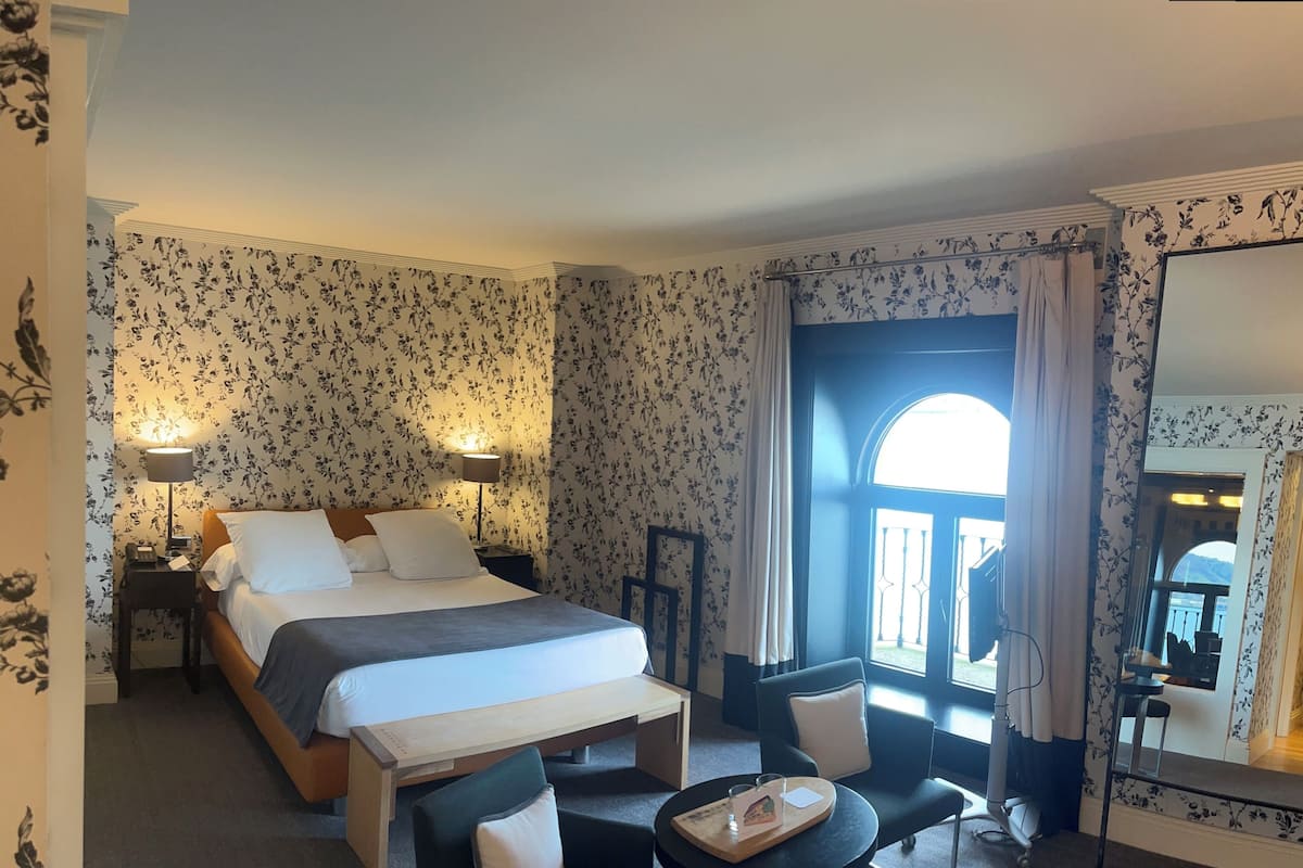A hotel room with a bed, bedside lamps, a desk with a chair, a small table with a tray, and a large mirror.