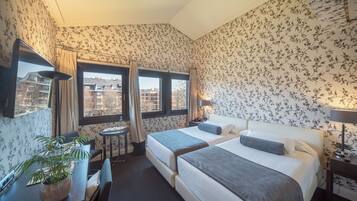 Habitación de hotel con papel tapiz floral, dos camas, un escritorio, una silla, una planta en maceta y vistas a edificios a través de las ventanas.