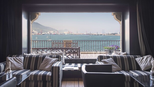 A balcony with striped sofas and a table, overlooking a harbor with boats and buildings.