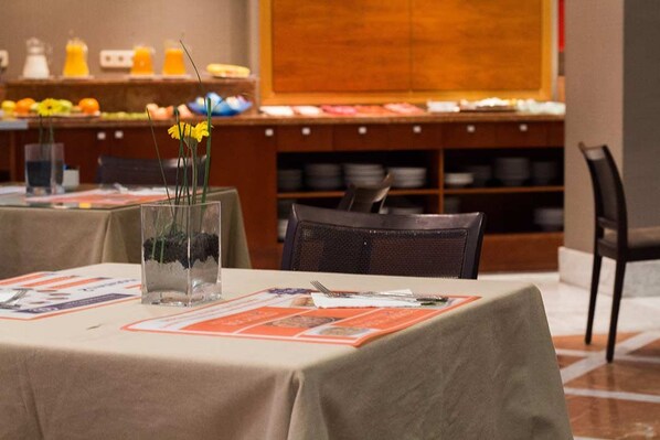 Uma área de refeições com mesas preparadas para hóspedes, um buffet com diversos pratos e um vaso decorativo com flores.