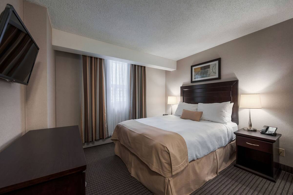 A hotel room with a bed, bedside lamps, a desk, a TV, and a painting on the wall.