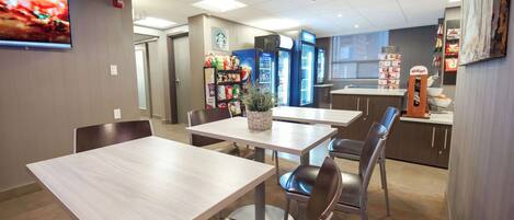 A dining area with tables and chairs, a television, vending machines, and a kitchen area with a refrigerator and shelves.