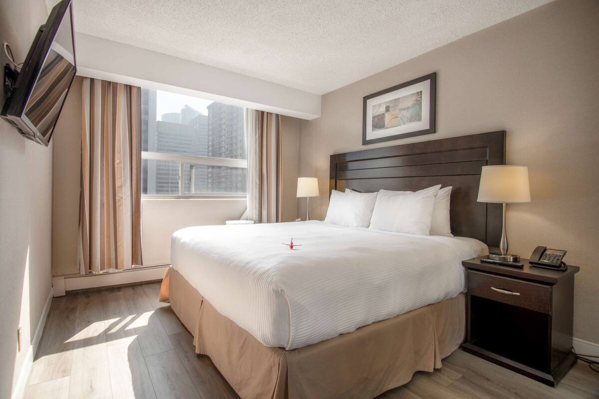 A hotel room with a large bed, two bedside tables with lamps, a flat-screen TV mounted on the wall, and a window with curtains.