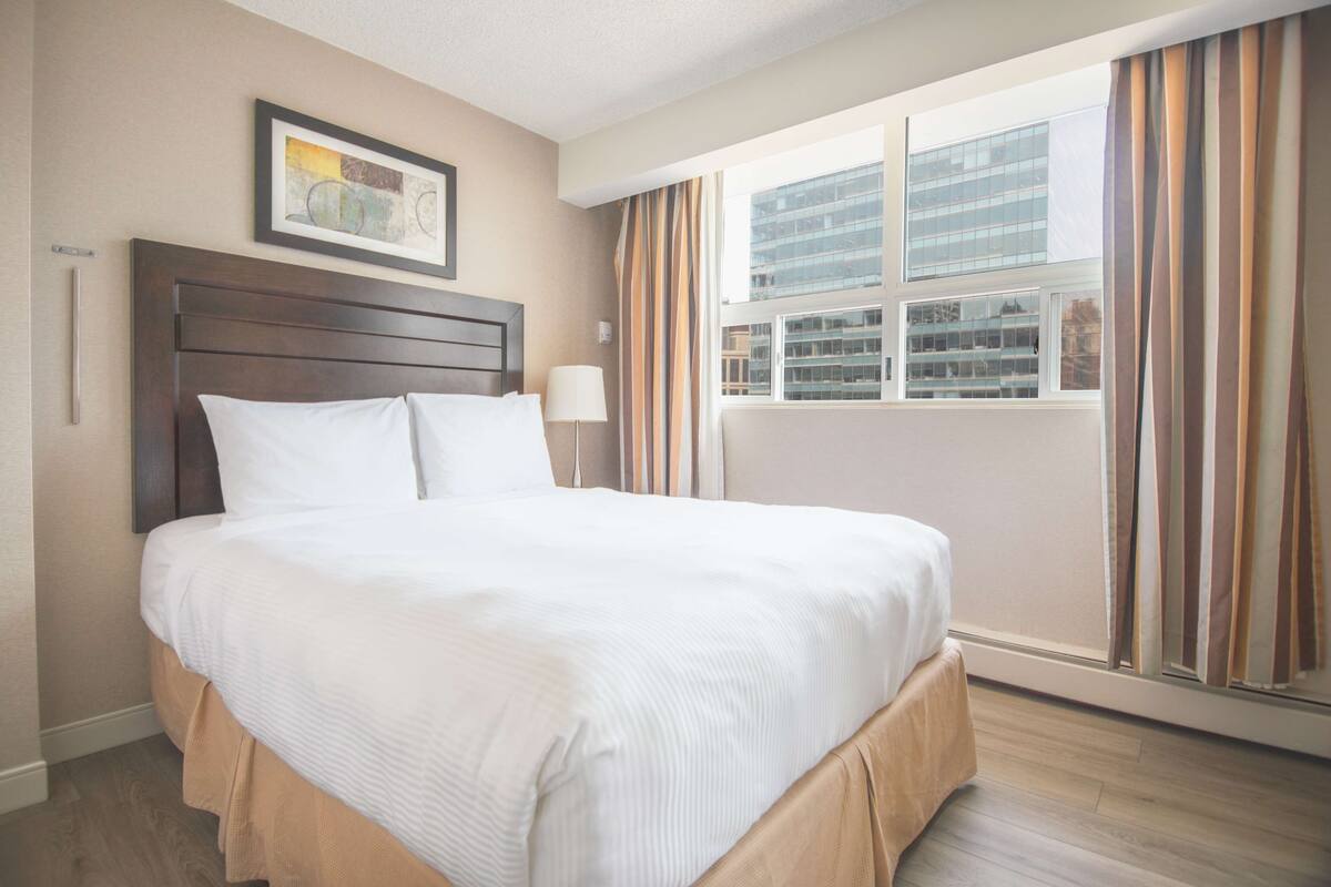 A hotel room with a large bed, a headboard, a window with curtains, and a framed picture on the wall.