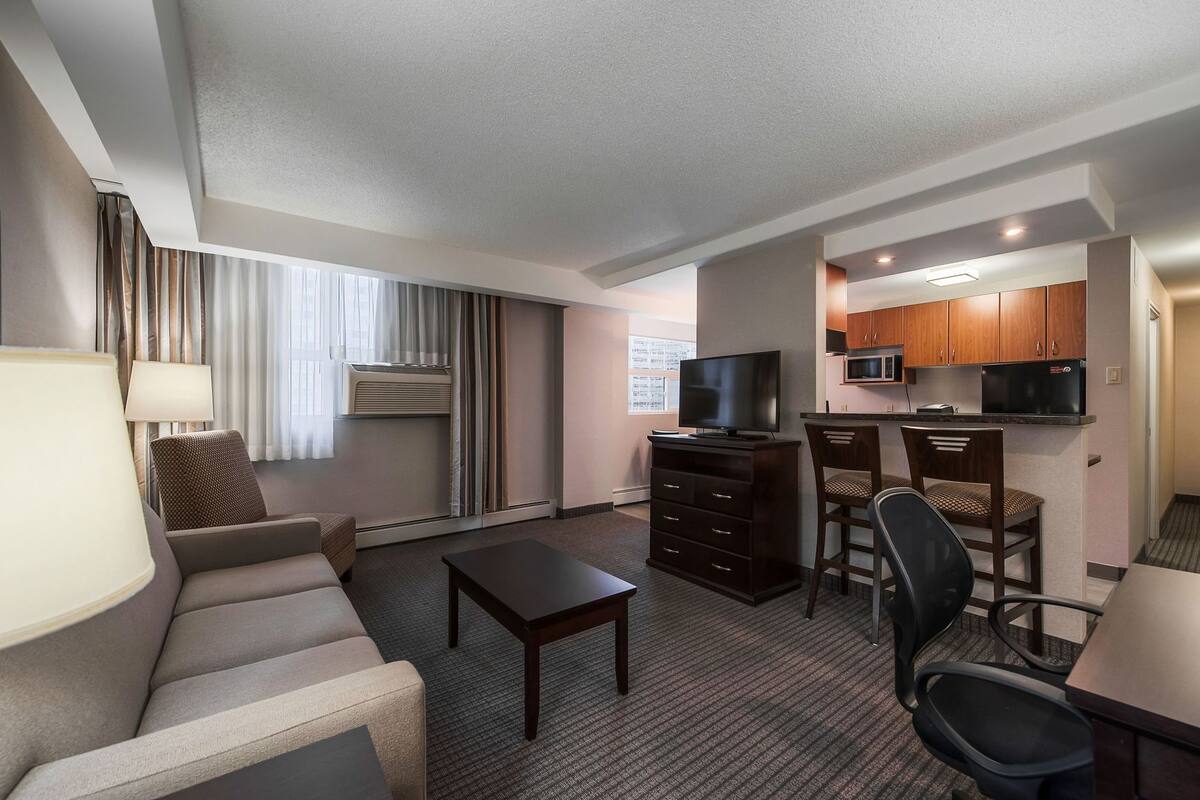 A hotel room with a sofa, a TV, a small table, a kitchenette, and a window with curtains.
